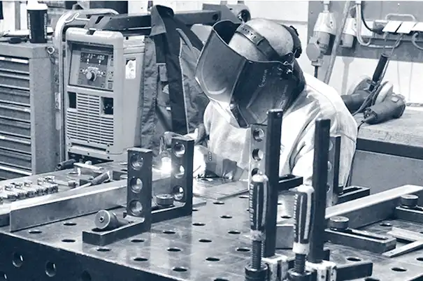Eine Person mit Schweißhelm arbeitet in einer Industriewerkstatt mit Metallteilen.